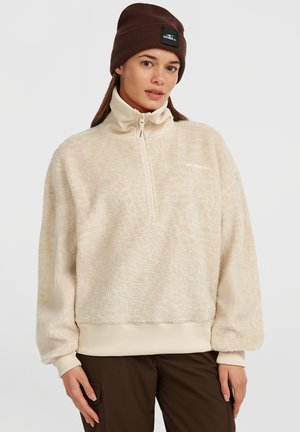 Kvinne som har på seg en beige fleece-genser med kvart glidelås og en brun lue, stående med avslappede hender mot en ensfarget bakgrunn.
