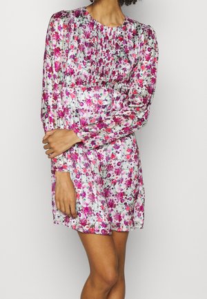 Kobieta w sukience z długim rękawem w kwiatowy wzór w kolorach różowym, fioletowym i czerwonym na jasnym tle, stojąca na tle gładkiej ściany.