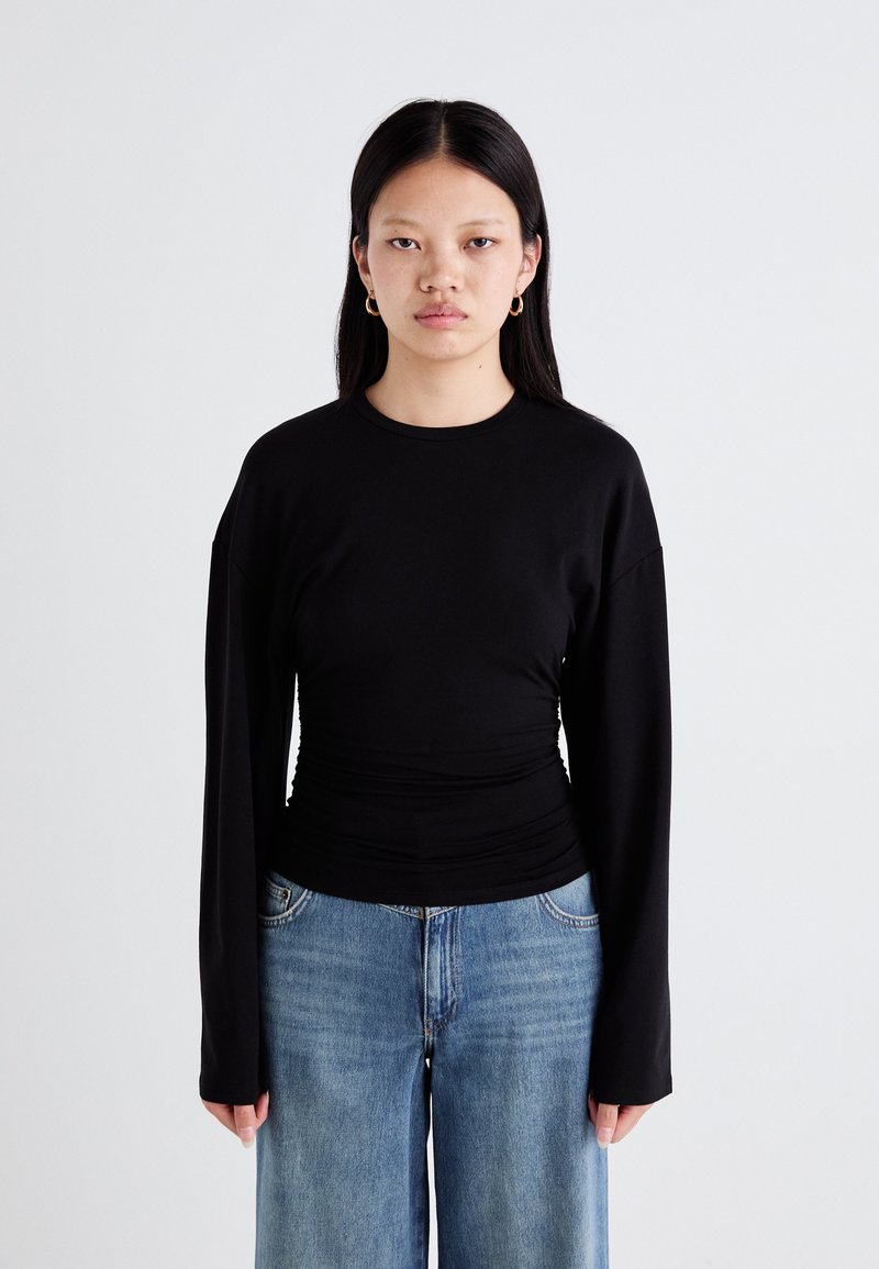 Black long-sleeve fitted top with gathered waist detailing, paired with light blue wide-leg jeans. Plain texture, no visible patterns or hardware.