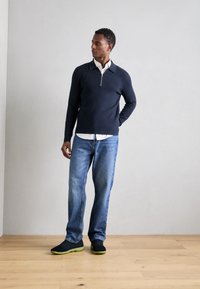 Felpa navy a mezza zip, camicia bianca con colletto sotto, jeans blu e scarpe nere con suole verdi. In piedi contro uno sfondo chiaro.