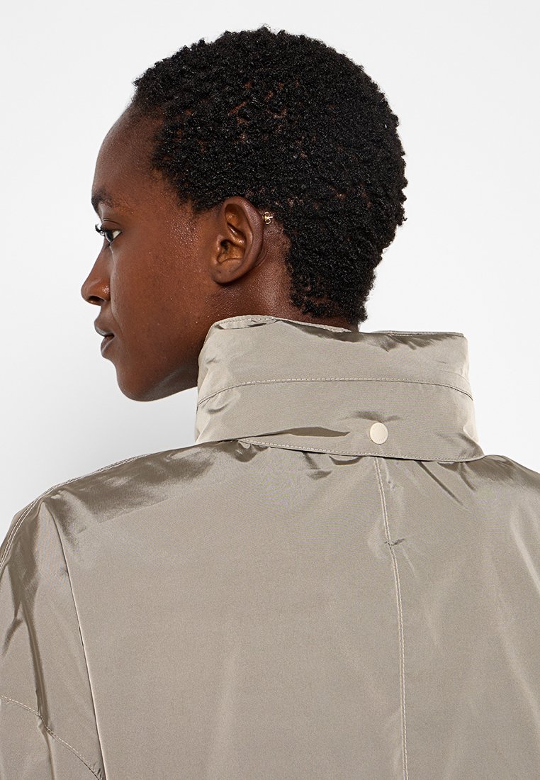 Giacca beige chiaro realizzata in tessuto lucido, con colletto alto, chiusura a bottoni a pressione e dettagli di cucitura discreti sul retro.
