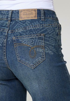 Denim kavbojke v temno modri barvi s zbledelim vzorcem. Imajo blagovno znamko usnjeni oznaki, značilno šivanje zadnjega žepa in kovinske zakovice.