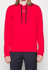 Czerwona bluza z kapturem wykonana z miękkiego materiału, posiada przednią kieszeń, czarne sznurki do ściągania oraz małą naszywkę z logo na piersi. Długie rękawy i ściągacze na mankietach.