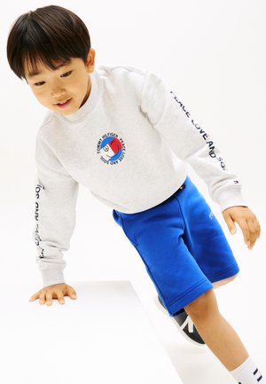 Ung dreng, der læner sig op ad en hvid overflade, iført en grå sweatshirt med cirkulært logo og blå shorts, ser fokuseret og afbalanceret ud.