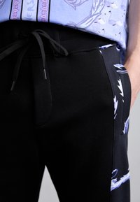 Melnas sporta bikses ar auklu viduklī, sānu kabatām un grafisku rakstu gar malām. Mīksts audums ar gludu tekstūru.
