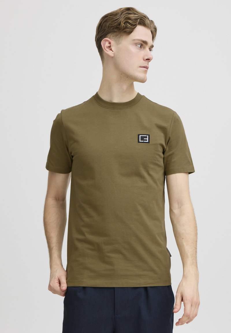 Tricou din bumbac de culoare verde măslin, cu guler rotund și mâneci scurte; are o mică etichetă cu logo alb-negru pe partea stângă a pieptului.