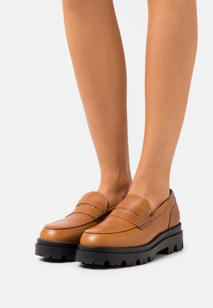 Mocassins en cuir marron avec un détail de sangle texturée et une semelle épaisse en caoutchouc noir, offrant une apparence chunky et robuste.