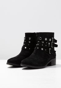 Botas de tobillo de ante negro con múltiples correas y tachuelas plateadas, cremallera lateral y un tacón bajo apilado. Textura suave y punta redondeada.