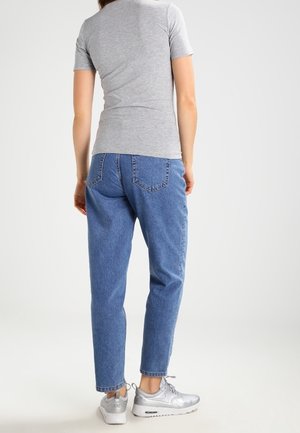 Femme portant un t-shirt gris à manches courtes, un jean taille haute bleu et des baskets argentées, debout de dos devant un fond uni.