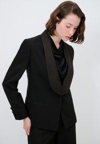 Musta frakki, jossa on satiininen kauluspanta, muotopuku ja napilliset hihansuut; yhdistetty mustaan satiiniseen toppiin tyylikkään ilmeen saavuttamiseksi.