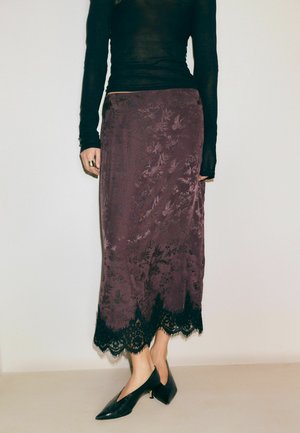 Žena má na sebe fialovú kvetovanú midi sukňu s čiernou čipkovanou lemovkou, čierne priliehavé tričko s dlhými rukávmi a čierne špicaté topánky s nízkym podpätkom, stojaca pri bielej stene.