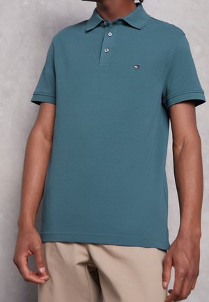 Man draagt een blauwgroen poloshirt met korte mouwen en een klein logo op de borst, gecombineerd met beige broek, staand tegen een grijze achtergrond.
