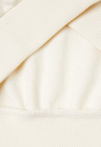 Krémová látka se hladkým, strukturovaným povrchem a žebrovanými akcenty. Vrstvy se překrývají a ukazují různé tkaniny a měkkosti.