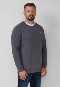Sweatshirt gris foncé avec un col rond, des manches longues et un petit logo sur la poitrine. Fabriqué à partir d'un tissu texturé et associé à un jean bleu.
