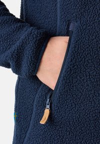 Navy fleece jacket with a zippered pocket, textured fabric, and contrasting zipper hardware. Features a small blue and yellow flag accent.