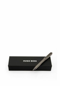 Stylo en métal avec une finition grise texturée repose sur une boîte noire étiquetée "HUGO BOSS" en lettres blanches. Le stylo possède un clip et une pointe lisse.