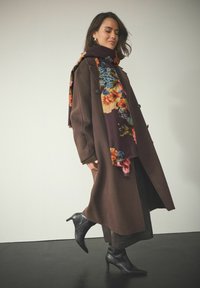 Brun uldfrakke med dobbeltknaplukning, parret med et flerfarvet blomsterhalstørklæde. Sorte hælhøje ankelsko fuldender outfittet.