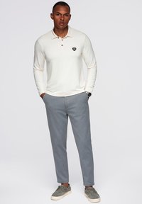 Polo a maniche lunghe in crema con collo a tre bottoni e logo sul petto, abbinato a pantaloni grigi chiari a righe e sneakers grigie.