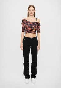 Kuttet, off-shoulder topp i oransje og svart dyreprint; kombinert med svarte jeans og hvite joggesko. Modellen står mot en nøytral bakgrunn.