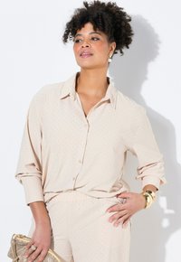Blusa beige con puntos, de mangas largas, con botones en la parte delantera y cuello puntiagudo. Tejido suave con una textura lisa; combinada con pantalones a juego.