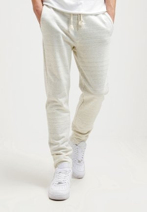 Homme portant un pantalon de jogging beige clair avec taille à cordon et des baskets blanches, debout les mains dans les poches sur un fond uni.