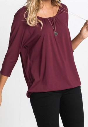 Femme portant un haut bordeaux ample à manches longues et un pantalon noir, tenant un collier pendentif d'une main.