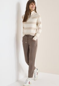 Femme debout portant un pull beige à rayures blanches, un pantalon marron à cordon de serrage et des baskets blanches avec des détails bordeaux sur un fond bicolore.