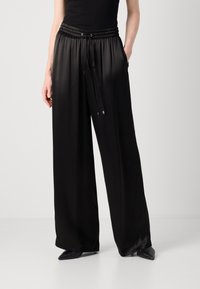 Una persona indossa un top senza maniche nero e pantaloni neri a gamba larga, con le mani in tasca, appoggiata a uno sfondo chiaro e semplice.