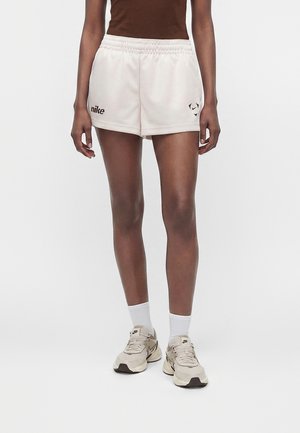 Osoba nosząca białe sportowe krótkie spodenki Nike, beżowe buty sportowe Nike, białe skarpetki oraz brązową bluzkę, stojąca na jednolitym tle.