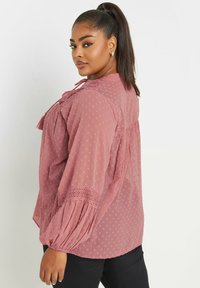 Blusa rosa con design a pois testurizzati, maniche arricciate e dettagli in pizzo, con un nastro sul retro. Pantaloni neri.
