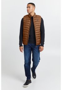 Braune gesteppte Weste mit hohem Kragen, getragen über einem dunklen Pullover. Kombiniert mit blauen Jeans und schwarzen Turnschuhen. Einfache Reißverschlussdetail.