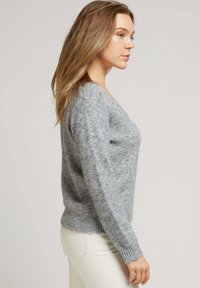 Pull en maille grise avec un col en V et des manches longues, doté d'une texture douce et de poignets côtelés, associé à un pantalon de couleur claire.