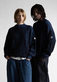 Du du jaunų suaugusiųjų, vilkintys derančius mėlynus ir juodus dryžuotus megztinius bei džinsus, stovi su rankomis kišenėse prieš paprastą foną.