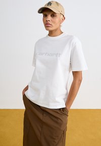 Fehér pamut póló világosszürke "carhartt" logóval, barna szoknyával párosítva. Bézs sapka sötét "LA" logóval.