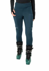 Skihosen in Dunkelblaugrün, aus dehnbarem Material mit hohem Bund. Mit seitlichen Taschen und reflektierenden Akzenten. Getragen mit Skischuhen.
