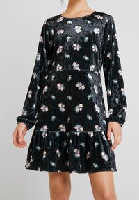 Svart sammetsklänning med långa ärmar, blommönster i rosa och grönt, volangdetaljer i nederkant, rund halsringning och åtsittande liv.