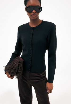 Žena v čiernom zapnutom kardigane, tmavých nohaviciach, s veľkými čiernymi slnečnými okuliarmi, nesie tmavohnedú kabelku s strapcami.