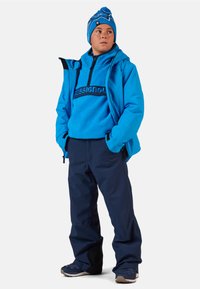 Sininen hiihtotakki hupulla, fleece-välikerros logolla, tummansiniset hiihtohousut ja yhteensopiva sininen pipo pompomin kanssa. Kestävä kangas ja käytännöllinen muotoilu.