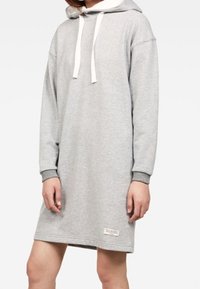 Grå huvtröjade klänning med långa ärmar, snörning i halsen och ribbade muddar. Har en etikett nära nederkanten.