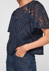 Top en dentelle bleu marine avec un motif de feuilles complexe, manches courtes et coupe décontractée, associé à un jean en denim foncé.