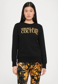 Čierna mikina s nápisom "VERSACE JEANS COUTURE" v zlatej farbe. Má okrúhly výstrih a rebrované manžety. Nosené s vzorovanými nohavicami v zlatej a čiernej farbe.