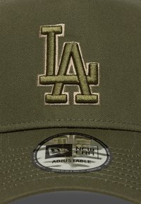 Oliven grønn caps med en brodert beige "LA" logo. Har en rund justerbar sticker på skjermen, laget av teksturert stoff.