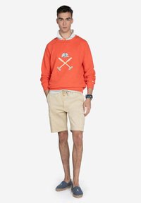 Sudadera naranja con un diseño gráfico en contraste, pantalones cortos beige y zapatos azul marino tipo slip-on. La tela parece suave; los pantalones cortos son de estilo cargo.