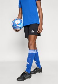 Μπλε αθλητική φανέλα με μαύρο σορτς, με λευκό λογότυπο Adidas. Μπλε κάλτσες με λευκές ρίγες και μαύρα παπούτσια ποδοσφαίρου. Κρατάει μια πολύχρωμη μπάλα ποδοσφαίρου.