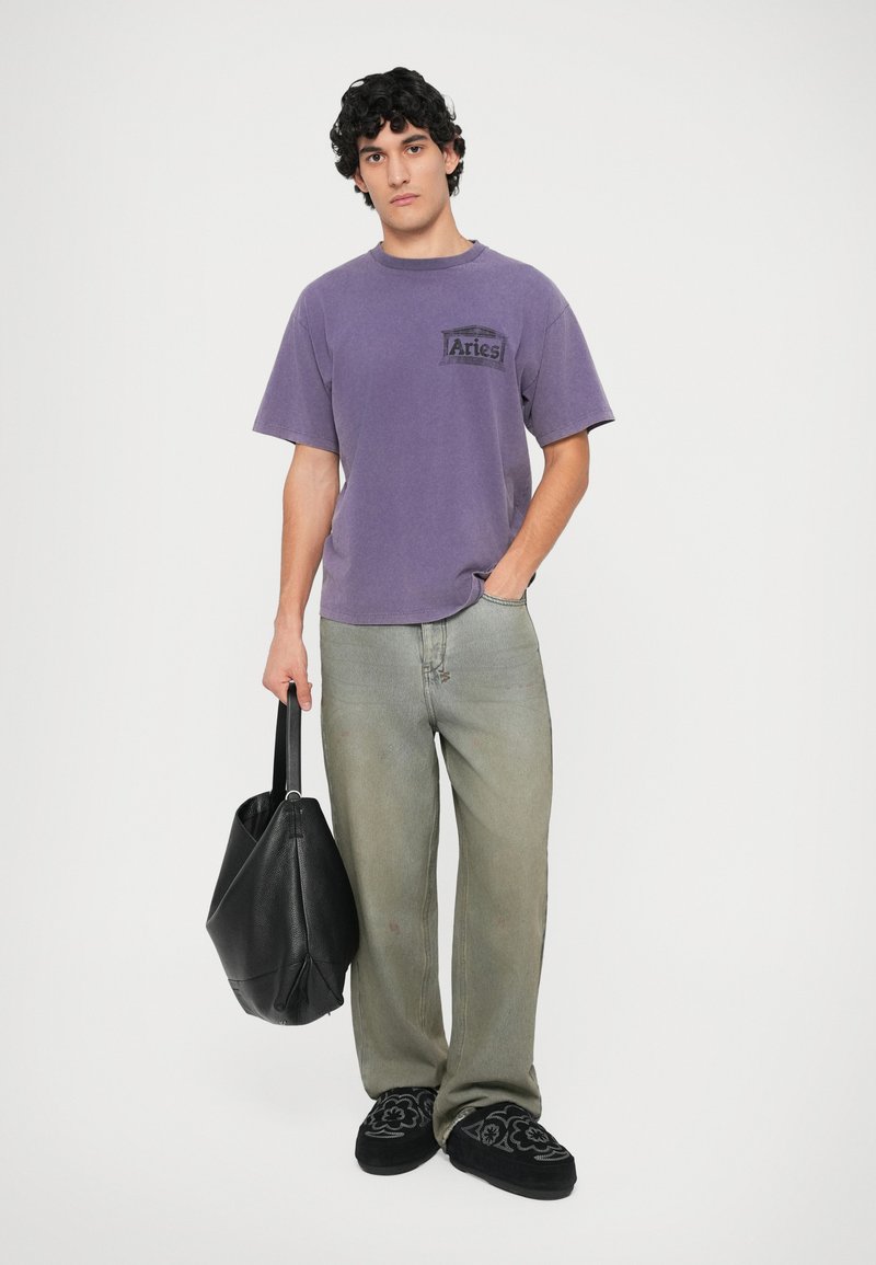 Un tricou din bumbac violet cu un logo Aries negru, combinat cu blugi largi gri deschis, o geantă neagră și pantofi slip-on negri brodați.