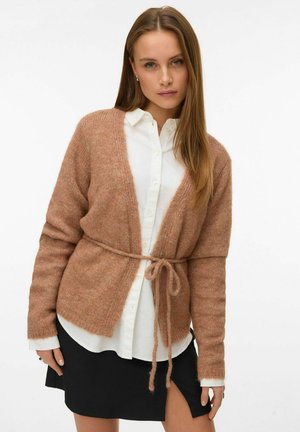 Cardigan lavorato a maglia marrone chiaro con chiusura a laccio, maniche lunghe e una texture morbida, indossato sopra una camicia bianca con bottoni e una gonna nera.