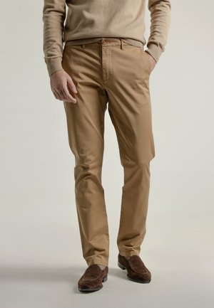 Hombre vestido con un suéter beige, pantalones chinos color beige y mocasines de ante marrón, con una mano en el bolsillo, frente a un fondo liso.