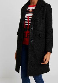 Manteau noir texturé avec un col cranté, des poches avant et deux boutons. Porté sur un pull festif rouge et blanc, avec un jean bleu.