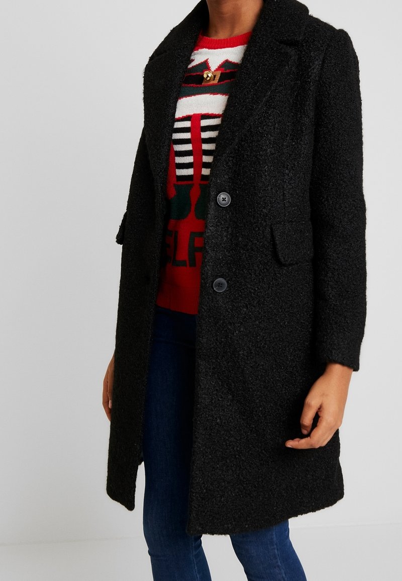 Manteau noir texturé avec un col cranté, des poches avant et deux boutons. Porté sur un pull festif rouge et blanc, avec un jean bleu.