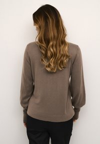 Brūns adīts džemperis ar garām, nedaudz pūkām piedurknēm un pieguļošu jostasvietu. Mīksta tekstūra un minimālistisks dizains.